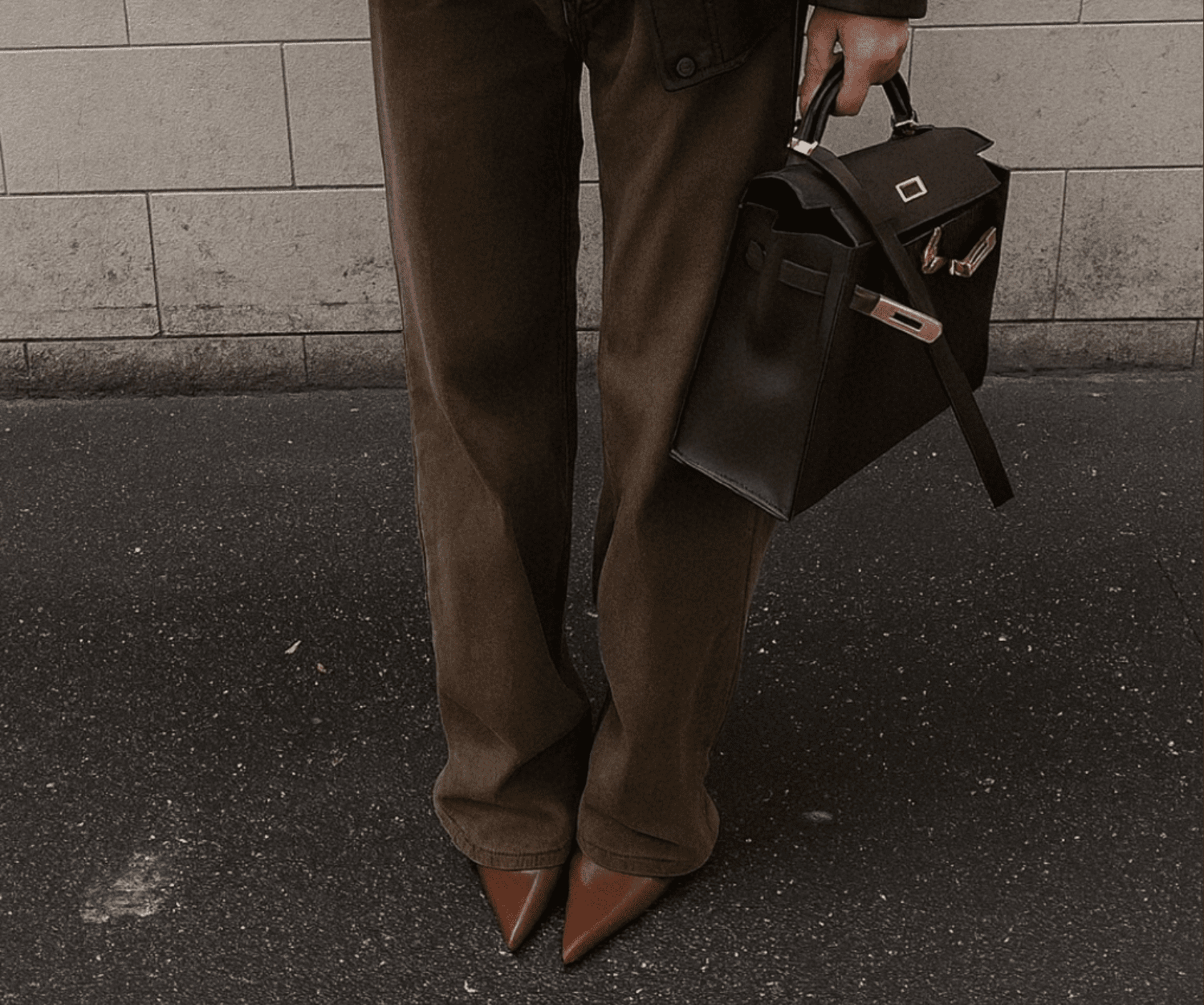 Woman in tailored dark trousers and pointed brown heels holding a black handbag on a city street