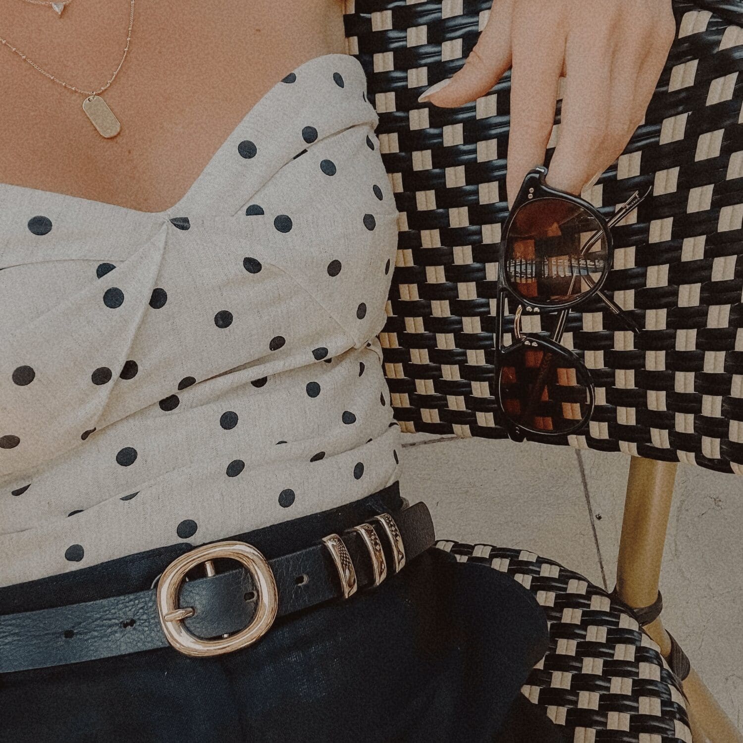 Close-up of a woman wearing a white polka dot blouse with a black belt, seated on a woven black-and-white café chair while holding a pair of sunglasses.